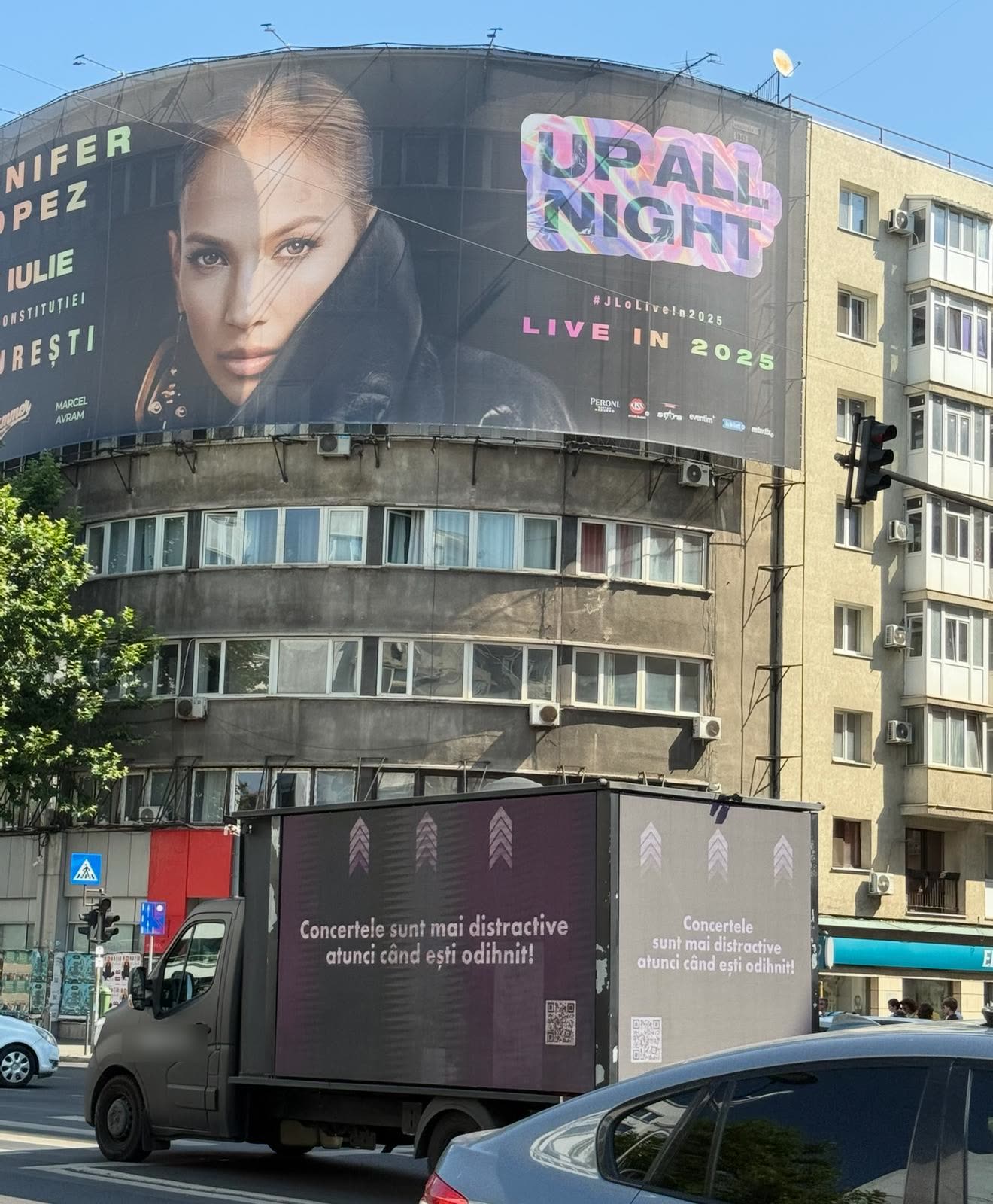 The van passing a building wrapped in a giant J.Lo 'Up All Night' banner, with the line 'Concertele sunt mai distractive atunci când ești odihnit!' (Concerts are more fun when you're rested!).